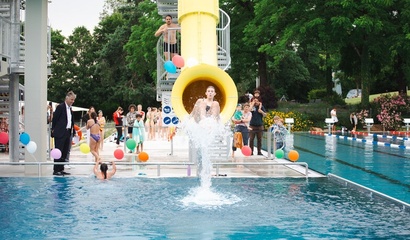 Ein Junge kommt aus einer gelben Wasserrutsche ins Schwimmbecken, umgeben von bunten Luftballons und Zuschauern am Beckenrand, anlässlich der Wiedereröffnung des Freibad Wyler..