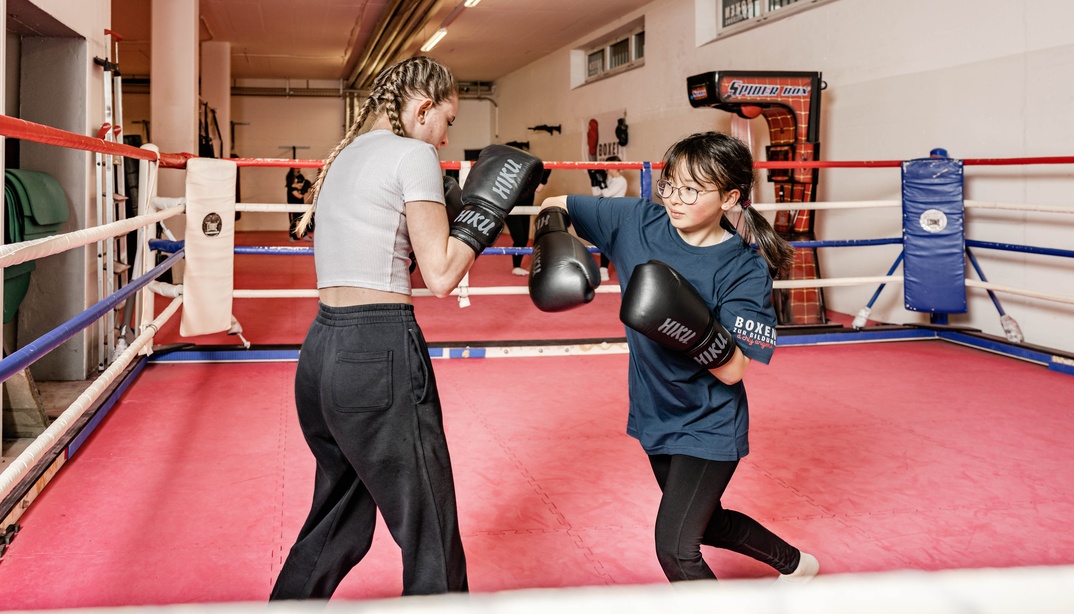 Zwei Mädchen mit Boxhandschuhen trainieren in einem Boxring; eine schlägt, die andere wehrt ab.