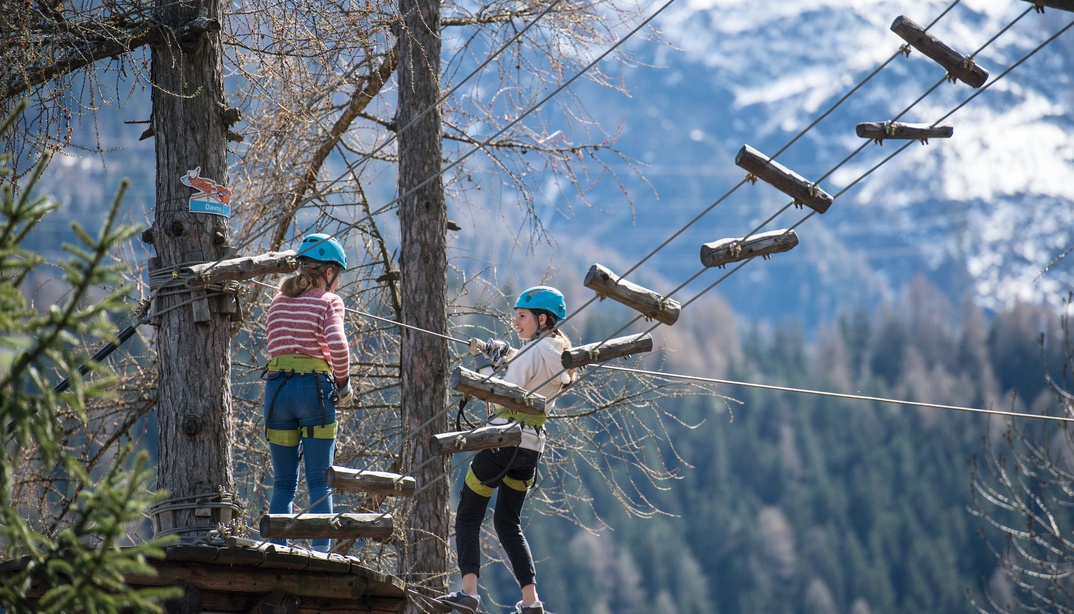 Zwei Kinder klettern auf einem Hochseilgarten-Parcours in den Bäumen, während sie Sicherheitshelme und Gurte tragen. Im Hintergrund sind Berge und Bäume zu sehen.