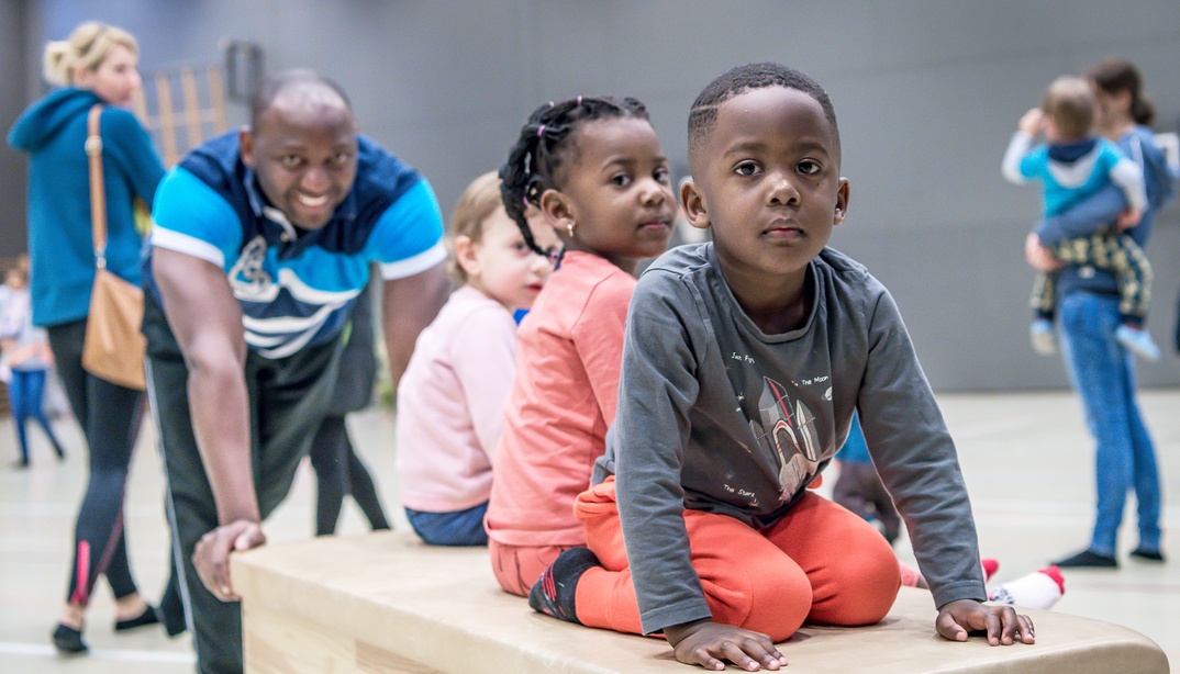 Kinder spielen in einer Halle und sitzen auf einem grossen Holzkasten, während Erwachsene in der Nähe sind.