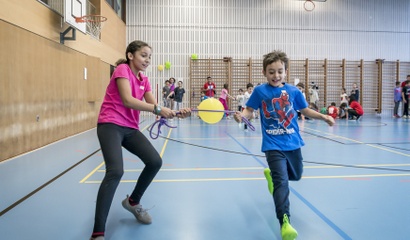 Zwei Kinder spielen in einer Turnhalle ein Teamspiel, bei dem sie gemeinsam einen gelben Luftballon mit einem Band halten und dabei laufen. Im Hintergrund sind weitere Kinder beim Spielen zu sehen.