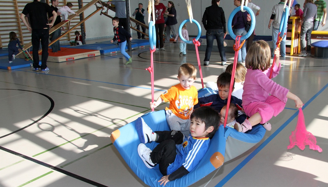 Mehrere Kinder sitzen auf einer grossen Mattel-Schaukel in einer Turnhalle.