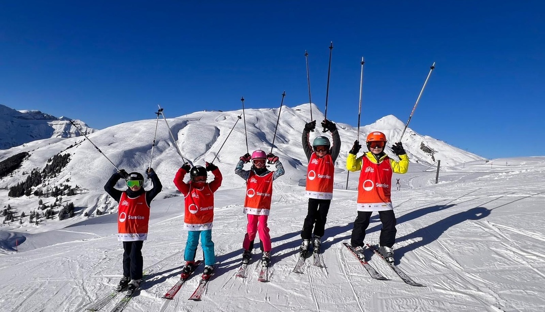 Eine Gruppe von Kindern in roten Skikleidung steht im Schnee und hält ihre Skistöcke hoch.