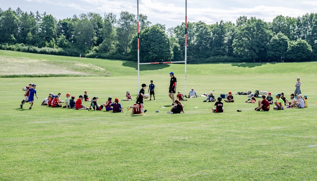 Eine Gruppe von Kindern sitzt auf einem weitläufigen, grünen Sportfeld unter freiem Himmel.