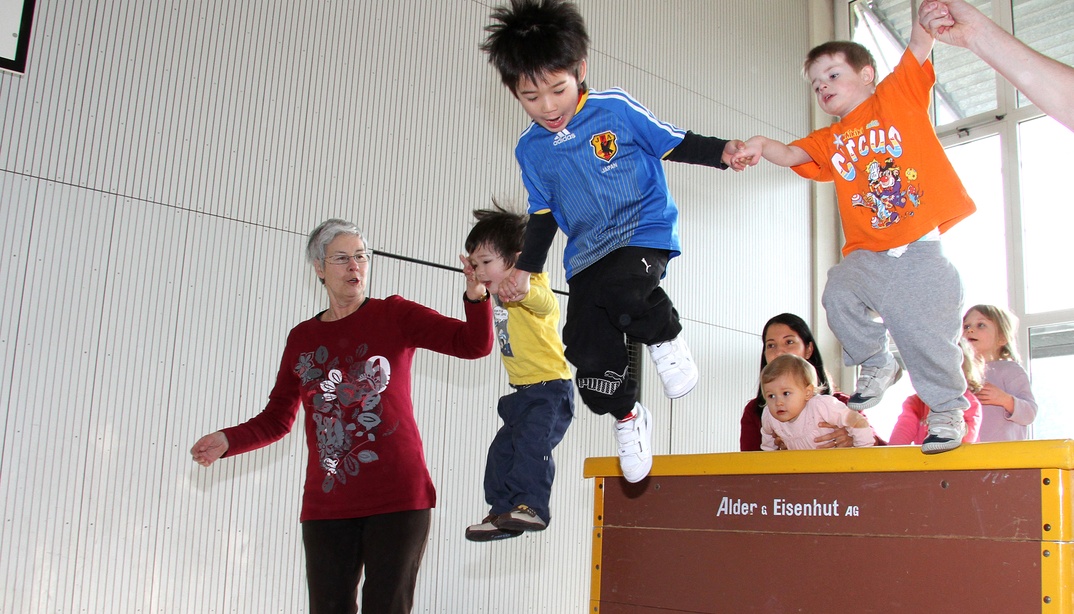 Kinder spielen und springen an der Hand von Erwachsenen von einem Kasten in einer Turnhalle