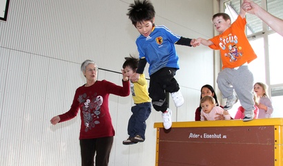 Kinder spielen und springen an der Hand von Erwachsenen von einem Kasten in einer Turnhalle