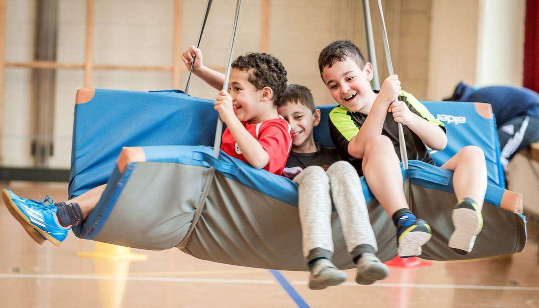 Drei Jungen spielen auf einer Matte an Seilen in einer Turnhalle