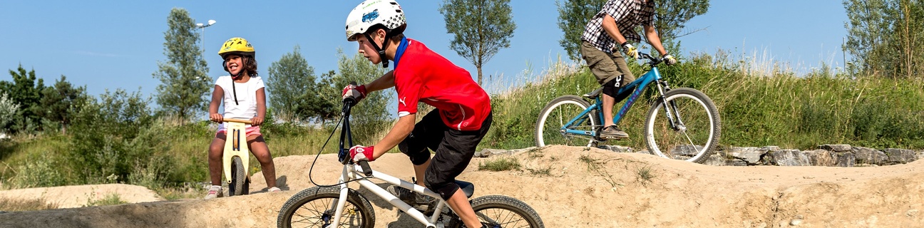 Zwei Kinder und ein erwachsener Mann fahren auf einem Pumptrack aus Erdmaterialien
